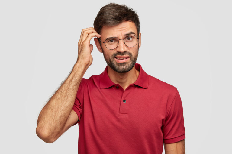 Puzzled unshaven guy scratches head, thinks deeply about something, has indignant look, dressed in casual clothes, stands against white background. Human facial expressions, emotions concept