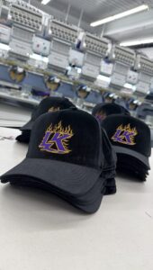 Stacks of custom embroidered black caps with a purple and gold flame logo in front of multi-head embroidery machines at Deerfield Custom Prints