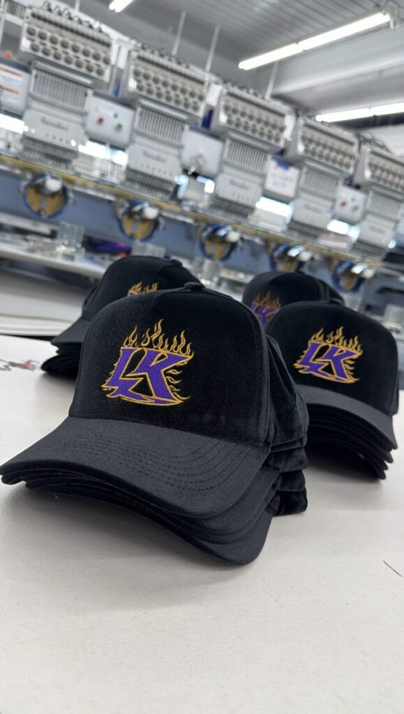 Stacks of custom embroidered black caps with a purple and gold flame logo in front of multi-head embroidery machines at Deerfield Custom Prints